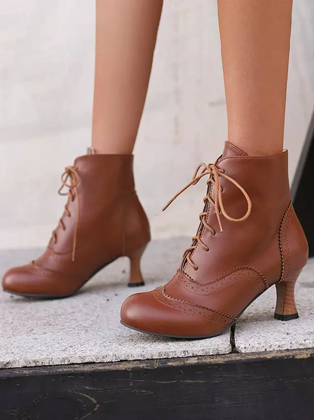 Brown vintage lace-up ankle boots with brogue details and curved brown mid-heel shown from side and frontal-side angle on outdoor concrete ground, displaying classic Victorian-inspired design elements