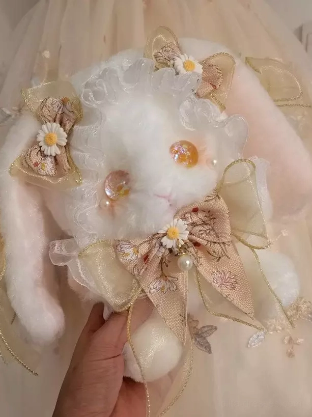 Plush bunny crossbody bag with brown printed bow and orange glass eyes, featuring ruffled lace trim, held by hand against soft fabric background