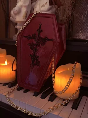 Dark gothic lolita coffin-shaped crossbody bag in burgundy with rose cross detail, three-quarter front view, piano keys background, gold metal chain strap detail