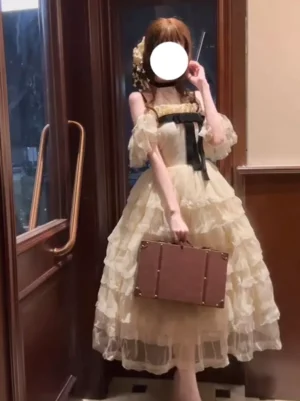 Off-shoulder champagne sweet lolita fairy dress with layered tuffle tulle skirt and front black bow, side profile on model holding vintage suitcase, puff sleeves design