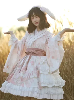 Cherry blossom sweet lolita one-piece dress in pink and white, featuring kimono collar, long bell sleeves, tiered ruffled skirt, pink waist bow, bunny ear bonnet, outdoor reed field setting, front full-length view