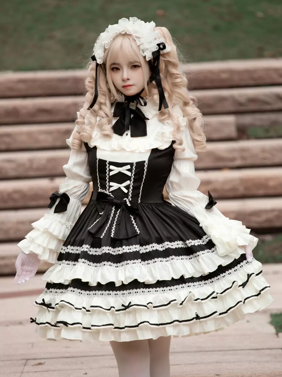 Black gothic lolita dress centered front view with crisscross lacing detail, bow necktie, and dramatic layered ruffled skirt on blonde model outdoor setting