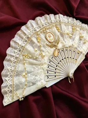 Cream white gold lace ruffle folding fan with pearl cameo ornament overhead view burgundy velvet background lolita accessory