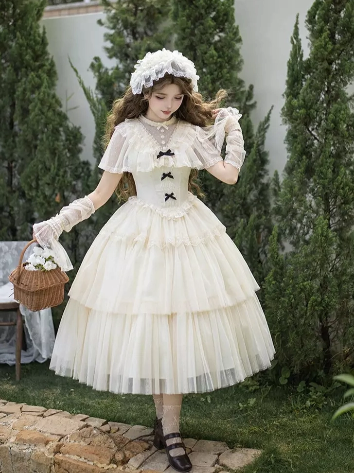 Left side angle of cream white Lolita dress set showing ruffled capelet, lace gloves, and dramatically layered tulle skirt on model with curly hair in garden setting