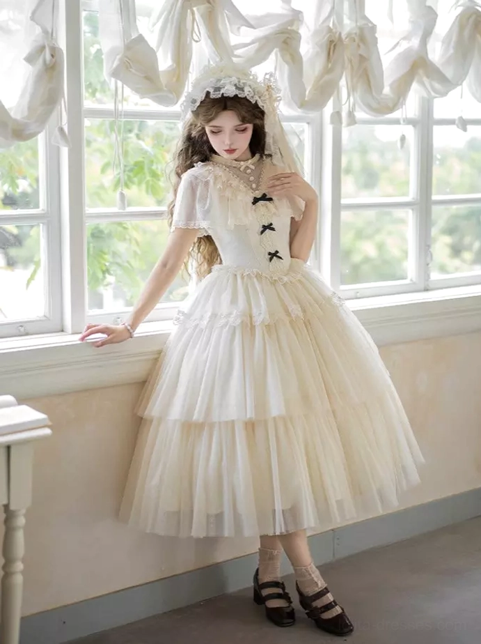 Three-quarter view of cream white Lolita dress with midi tiered tulle skirt, stand collar with bow details, and sheer cape near indoor window with model leaning on windowsill