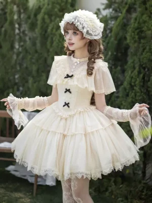 Cream white sweet lolita OP dress set front three-quarter view with sheer ruffled capelet, tiered tulle skirt, black bow details, and coordinating lace headdress in outdoor garden setting