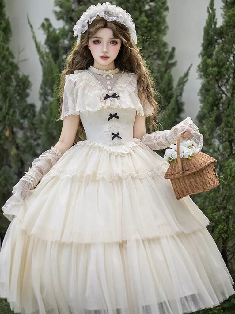 Cream white sweet Lolita one-piece dress with tiered tulle skirt, sheer cape collar, and black bow embellishments worn by model in outdoor garden setting with long curly hair