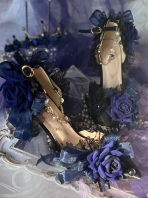 Deep blue lolita high heel sandals with rose and bow trims, pearl ankle chains, and pointed toe, displayed on vintage retro tray with purple tulle and miniature crown prop in three-quarter view