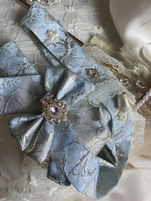 Dusty blue jacquard floral bow accessory with rhinestone and pearl embellishments and delicate lace trim, top-down close-up detailed view on white embroidered textile