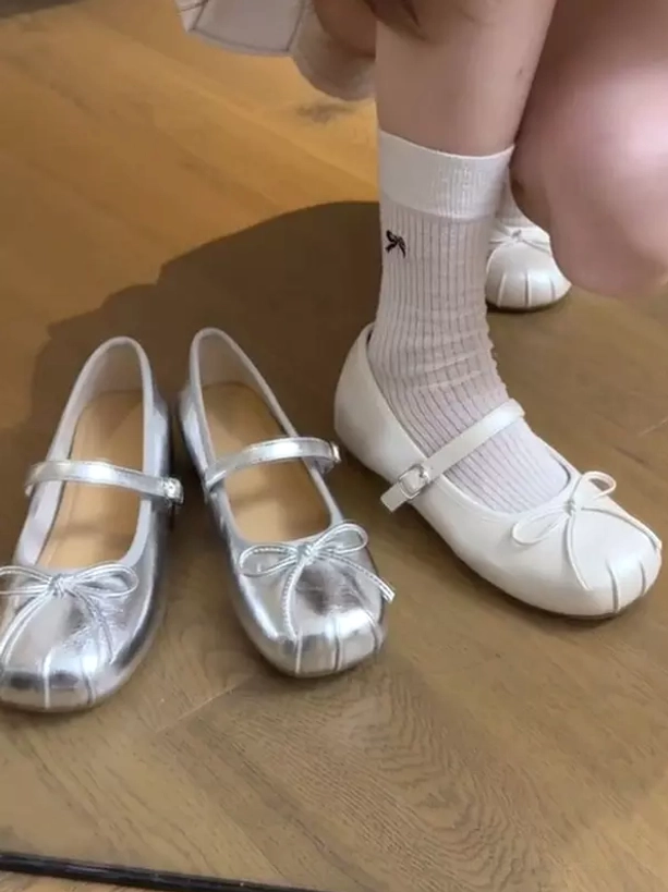 Ecru white ballet Mary Jane flat lolita shoes front overhead view with female leg wearing white ribbed socks, showing front bow embellishment and adjustable ankle buckle strap, matching silver color option displayed on wooden floor