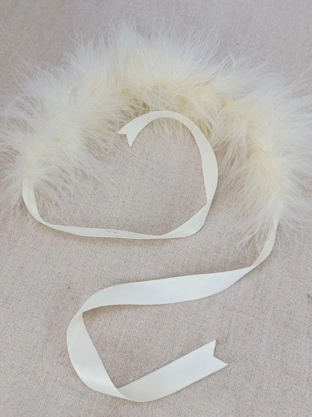 Close-up detail of fluffy ecru feather trim and satin tying straps of Lolita headdress, showing texture and craftsmanship of feather decor and ribbon components in flat lay view