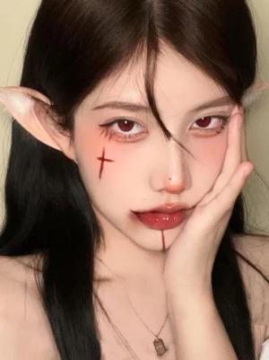 Enchanted elf ear cosplay accessory worn by model with gothic makeup and red contact lenses, frontal close-up portrait view showcasing light tan ear construction with white fuzzy pom-pom decorations