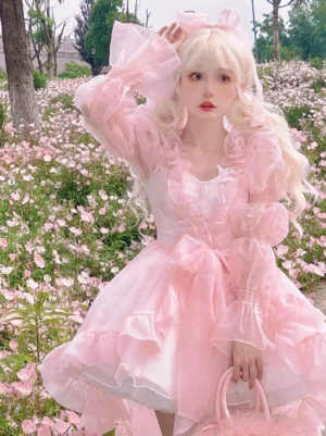 Enchanted Fairy sweet lolita OP dress front view featuring pink tiered ruffled skirt with decorative bow, sheer puff sleeves, and ruffled neckline on blonde model in flower field setting