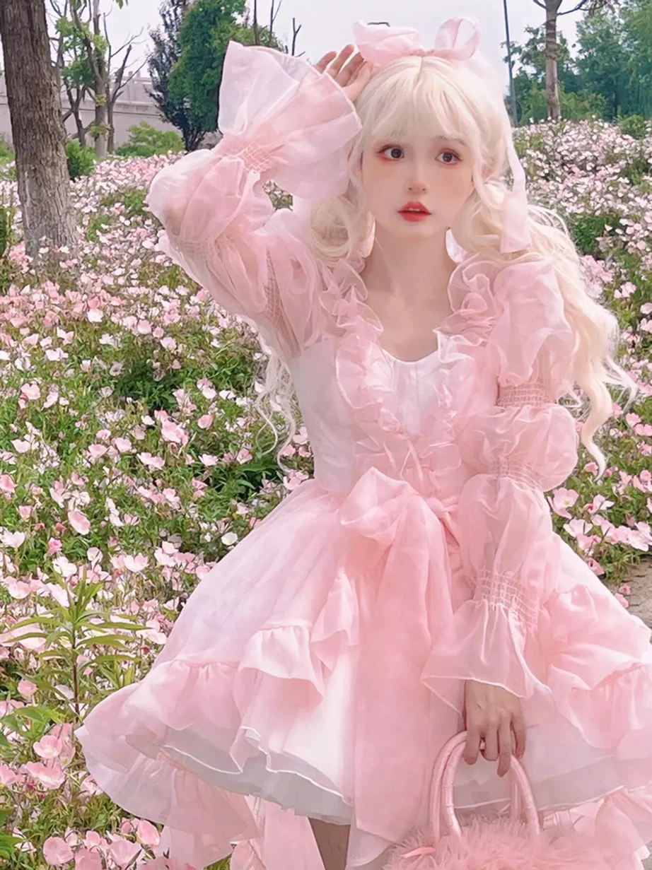 Enchanted Fairy sweet lolita OP dress front view featuring pink tiered ruffled skirt with decorative bow, sheer puff sleeves, and ruffled neckline on blonde model in flower field setting