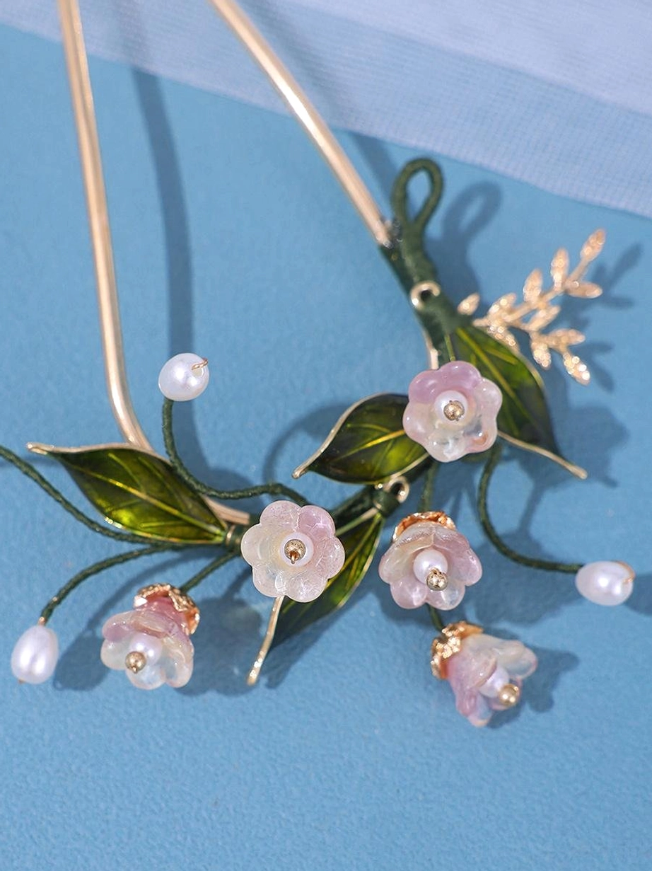 Close-up detailed view of curved flower hairpin showing intricate pink resin flowers, realistic green leaves and faux pearls on elegant gold metal structure against blue fabric backdrop