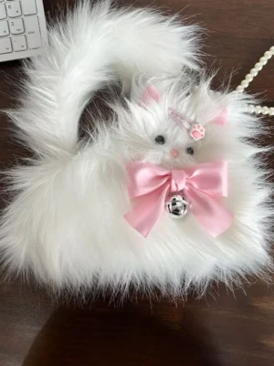 Top-down flat lay of plush white cat crossbody bag on dark desk showing detachable pearl chain strap, pink bow with bell, and overall bag structure