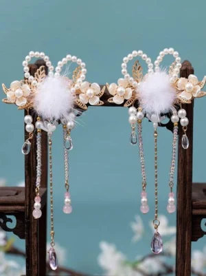Pair of gold vintage lolita hair clips with white pom-poms, pearl flowers, and beaded tassels displayed on wooden stand against teal backdrop, frontal view