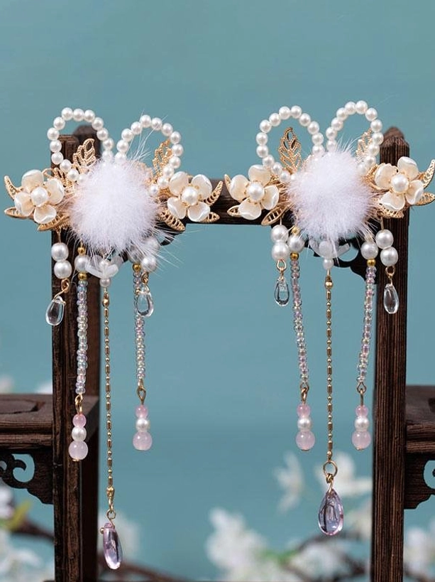 Pair of gold vintage lolita hair clips with white pom-poms, pearl flowers, and beaded tassels displayed on wooden stand against teal backdrop, frontal view