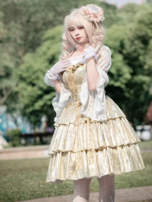 Golden tiered sweet lolita JSK dress with layered ruffle hemline worn outdoors on grass, featuring shiny golden polyester fabric and white lace accents, front three-quarter view