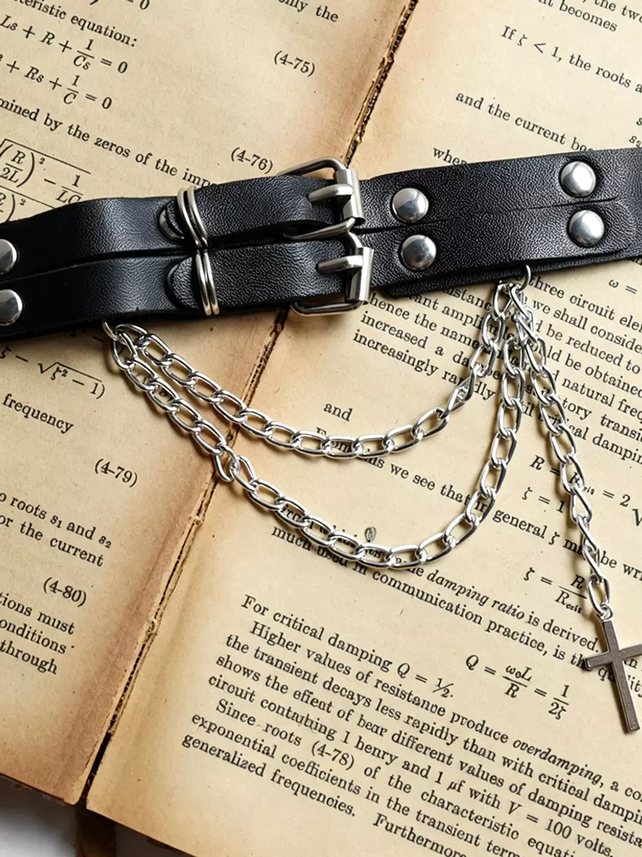 Gothic leather arm band close-up flat lay on vintage textbook pages showcasing double buckle system, silver rivets, chains and cross pendant design detail