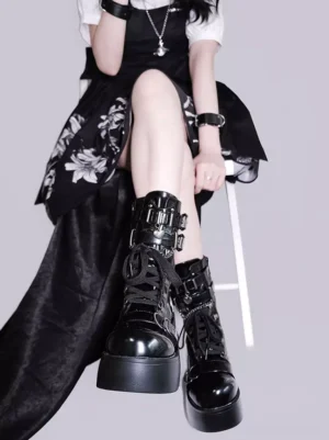Black platform combat boots with metal buckles and chain accents worn by gothic lolita model sitting on white stool, thick glossy patent leather chunky platform sole
