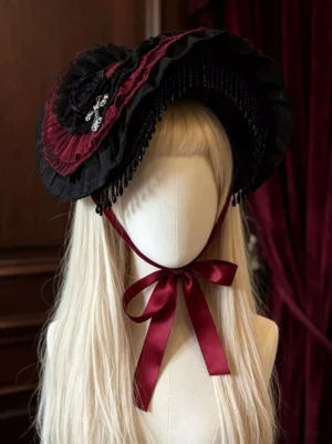 Gothic burgundy rose lace fascinator bonnet dark Lolita headdress front three-quarter view on mannequin with blonde wig, featuring black and wine red lace, beaded tassel fringe, cross ornament, and velvet rose decoration against dark wood and wine red velvet background