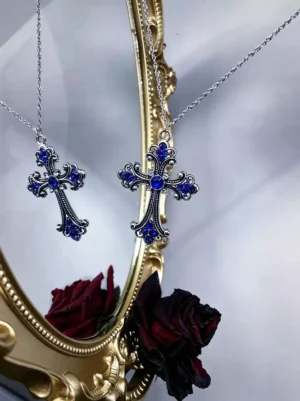 Ornate silver cross pendant with sapphire blue rhinestone inlays showcasing gothic lolita aesthetic on reflective gold mirror surrounded by romantic dried rose arrangement, detailed carved embellishments