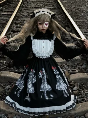 Gothic doll print sleeveless black vintage lolita JSK dress frontal view worn by blonde model with maid headband on railway tracks showing white ruffled lace trim and black-white doll pattern