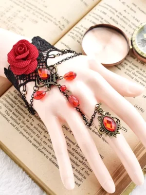 Gothic black lace hand bracelet with ring chain featuring red rose and red water-drop gem tassel, antique bronze metal details, oblique angle on vintage book background
