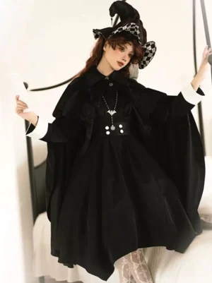 Black Gothic Lolita sleeveless cloak cape with layered ruffle collar worn by model in bedroom setting with witch hat, showcasing contrast white cuffs and waist button embellishments