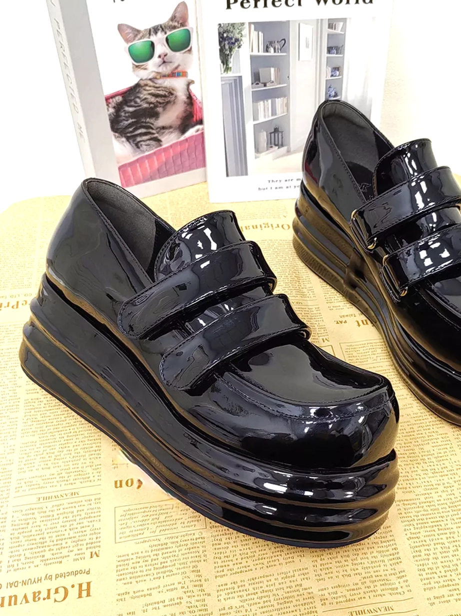 Three-quarter side view of black patent leather Lolita platform shoes pair showing layered wedge sole and hook-and-loop fastening with decorative themed backdrop