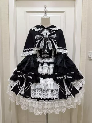 Gothic Lolita sleeveless black dress with oversized bow and detachable cape front view on dress form with white door background multi-layer lace trims pearl chain accents