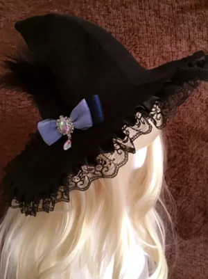 Gothic lolita black witch hat with lace ruffled brim and blue crystal bow, left oblique overhead view on blonde mannequin
