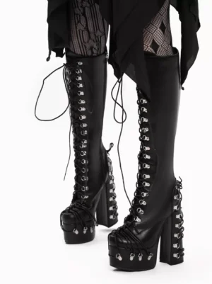 Black platform lace-up boots side-front three-quarter angle showcasing tall boot shaft, silver heel hardware, and trailing decorative laces with model in fishnet tights