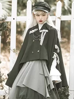 Black gothic lolita cape full front view with double-breasted buttons and bow embellishments paired with white ruffled blouse and picket fence outdoor setting