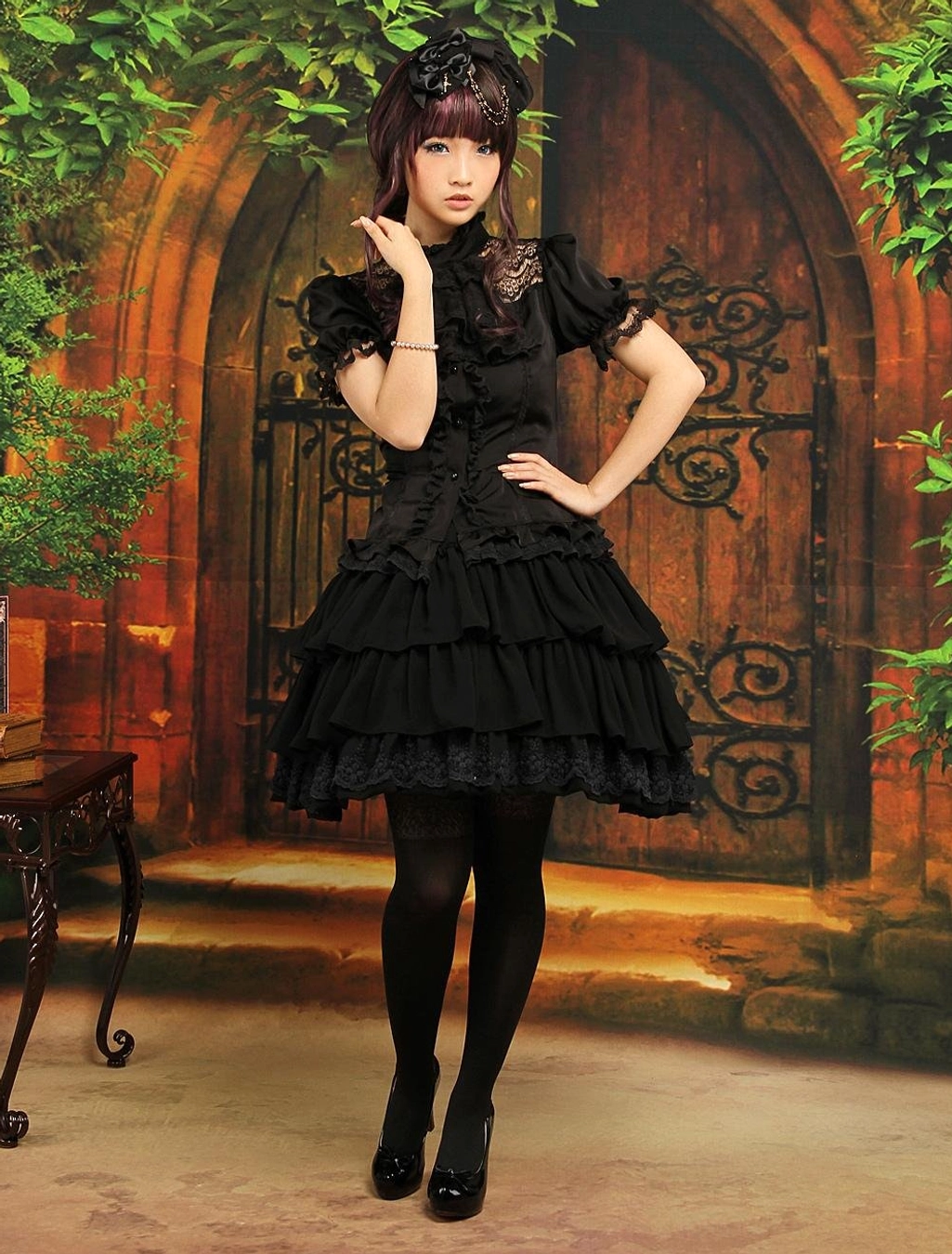 Gothic lolita coordinate set with black chiffon ruffled skirt worn with lace-collar blouse black tights and flat shoes full body standing pose vintage courtyard backdrop