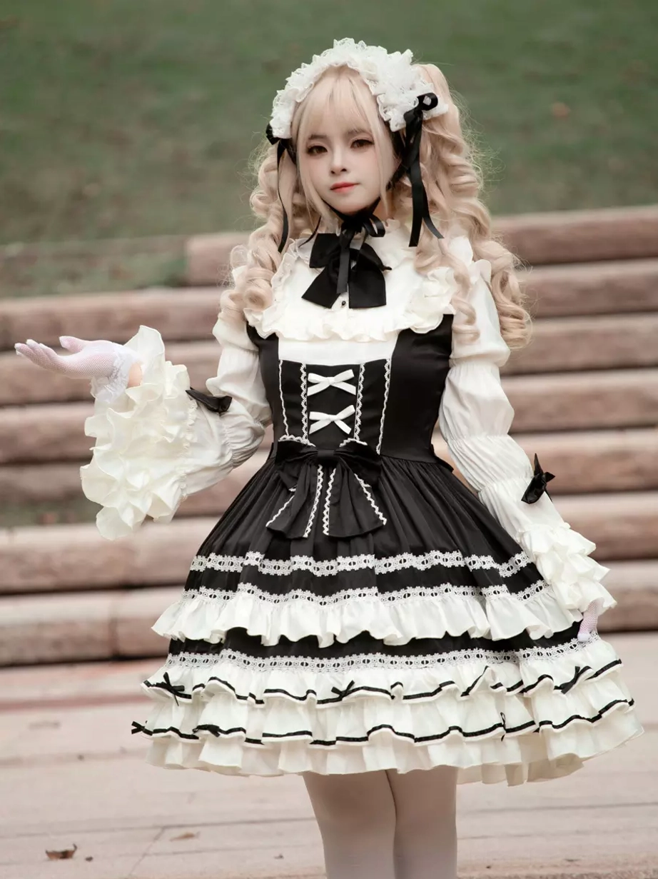 Classic black gothic lolita dress front full-body view with blonde model wearing long ruffled sleeves, black and white layered skirt, and decorative bow necktie on outdoor stone stairs