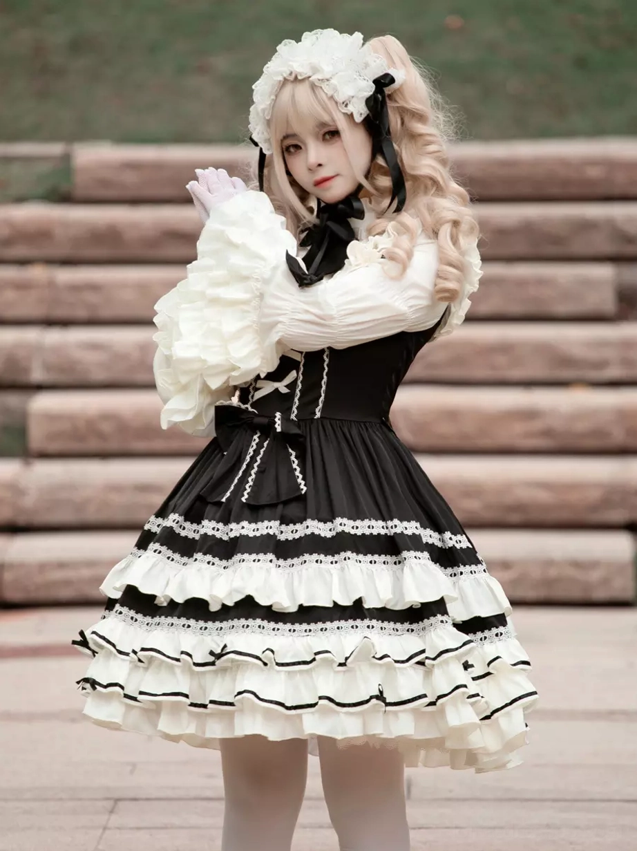 Black gothic lolita dress side-front partial view with model showing front chest bow decoration, clasped hands pose, and layered skirt construction