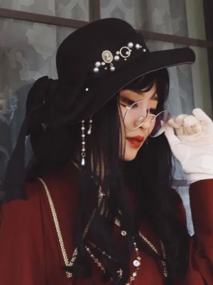 Gothic Lolita black wide-brim hat styled on female model wearing vintage red outfit, three-quarter front view showing pearl details, chain embellishments, and coordination with round glasses and white lace gloves