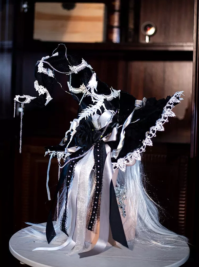 Right side three-quarter view of gothic Lolita witch hat accessory with black-white contrast, floral lace edging, hanging ribbon tassels, and silver decorative chains on dark wood bookshelf background