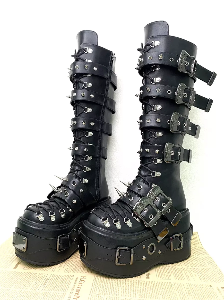 Black Gothic Lolita boots front three-quarter view showing carved decorative buckles, spike studs, lace-up design, and thick wedge platform sole on newspaper backdrop