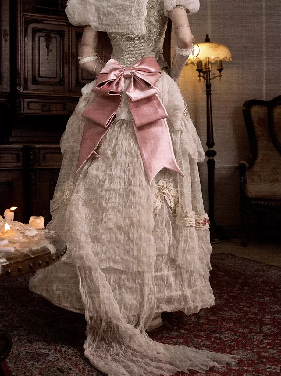 Back view of romantic pink satin bow worn as dramatic train decoration on cream ruffled lace vintage-inspired wedding dress with long skirt train in dim antique living room setting