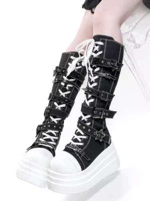 Black canvas gothic lolita platform boots with white chunky sole, star-shaped metal buckles, and lace-up front closure shown on model wearing dark outfit against white chair background