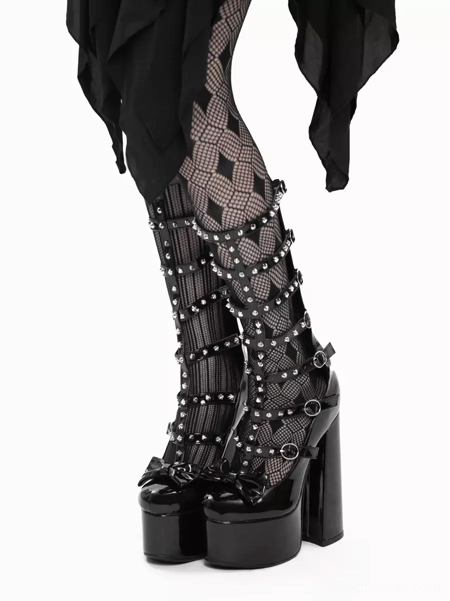 Front low-angle view of Gothic Lolita platform boots with studded buckle straps, patent leather platform, bow detail and wide block heel worn over diamond fishnet stockings and black draped skirt