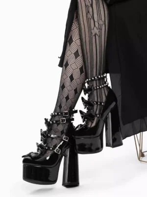 Side view of Gothic Lolita platform sandals featuring metallic glossy black patent leather, studded details, bow buckles, and thick platform heel, model wearing argyle fishnet stockings and black skirt seated on metal chair
