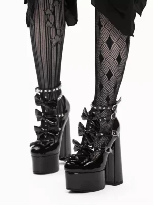 Gothic Lolita platform high heel sandals with glossy black patent leather, rivet ankle straps, bow decorations, and chunky block heel worn with argyle fishnet stockings and black irregular skirt - front close-up view