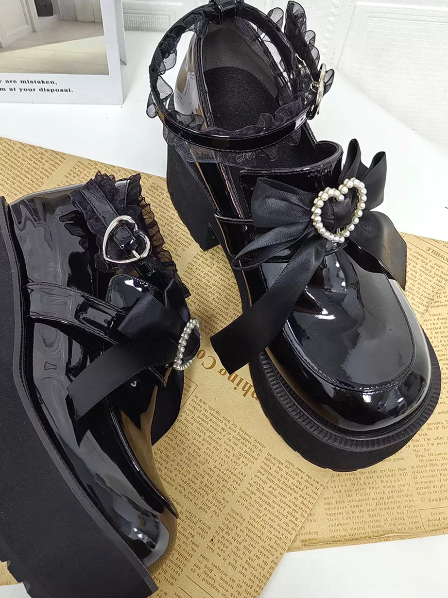 Overhead mixed side view of black gothic lolita platform pumps placed on vintage newspaper prop, featuring pearl-embellished heart bow decor and lace ankle trim detail