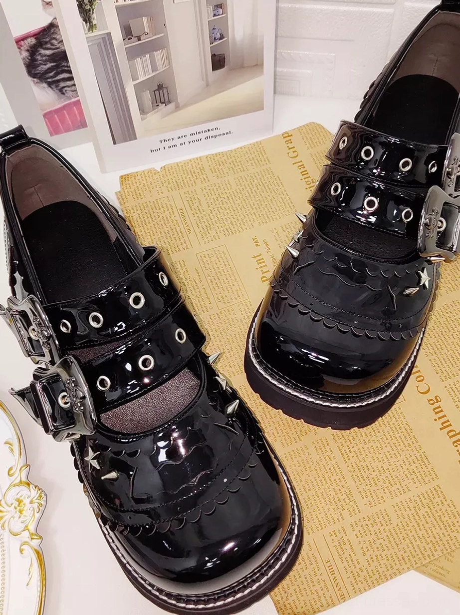 Black glossy gothic lolita platform shoes with double buckle straps and metal spike accents, arranged on tabletop with vintage newspaper and decorative artwork props, showing thick chunky heel construction and patent PU leather finish from oblique angle