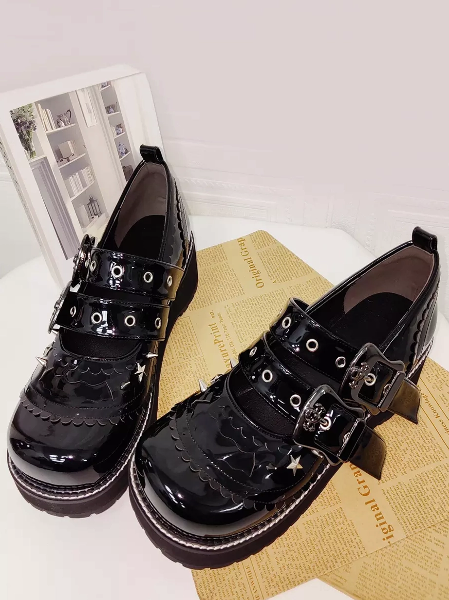 Pair of black gothic lolita pumps photographed from oblique top-down angle on vintage newspaper prop surface, showing star and spike metal decorations, dual adjustable buckles, and scalloped trim detail on black patent leather