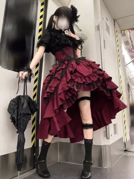 Gothic lolita two-piece set side full body view in subway carriage setting featuring high-low ruffled wine-red skirt, black short ruffled blouse, leg garter, and fashion umbrella prop